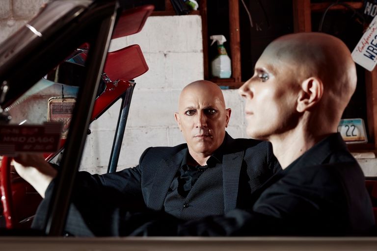 Rock band A Perfect Circle members Maynard James Keenan and Billy Howerdel wearing dark suits inside a classic car in a garage portrait.